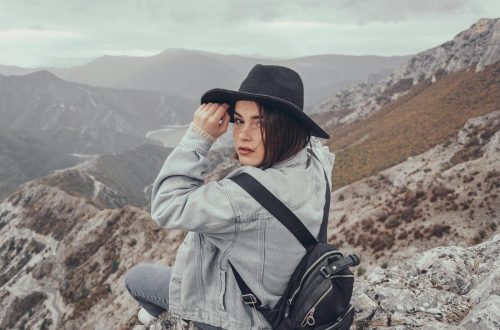 Back view of young female traveler in denim clothes and hat with backpack sitting on rocky mountain peak and looking at camera over shoulder