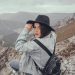 Back view of young female traveler in denim clothes and hat with backpack sitting on rocky mountain peak and looking at camera over shoulder